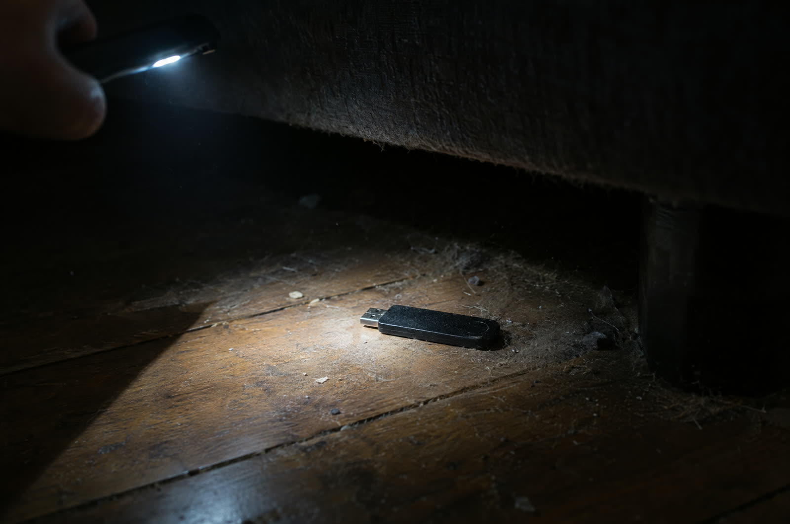 소파 밑 어두운 바닥에 떨어진 USB 지갑을 손전등으로 찾는 장면